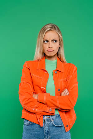offended woman frowning and pouting lips while standing with crossed arms isolated on green