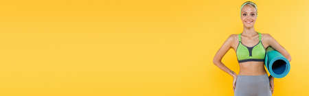 slim woman smiling at camera while standing with hands on hips and fitness mat isolated on yellow,...