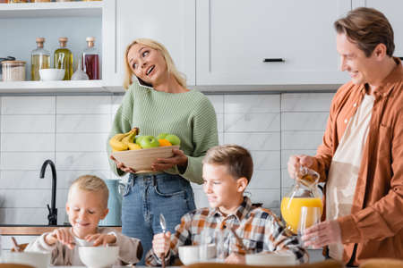 woman with bowl of fresh fruits talking on smartphone near man with orange juice ands boys having breakfastの写真素材