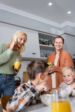 woman holding glass of orange juice and talking on smartphone near family having breakfast in kitchenの写真素材