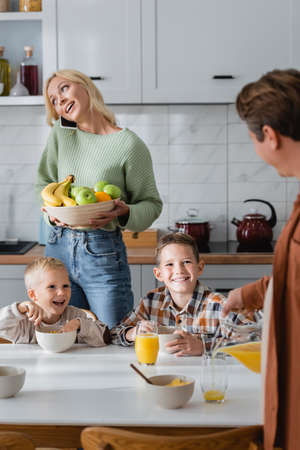 woman with fruits talking on mobile phone and man pouring orange juice near boys having breakfast in kitchenの写真素材