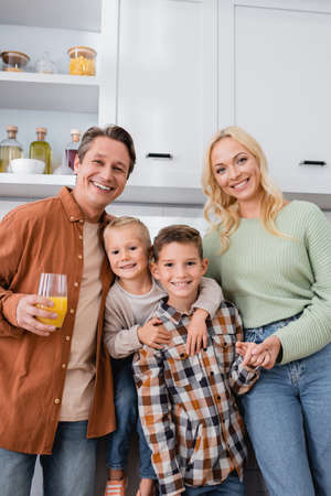happy man with orange juice smiling at camera near wife and sons embracing in kitchenの写真素材