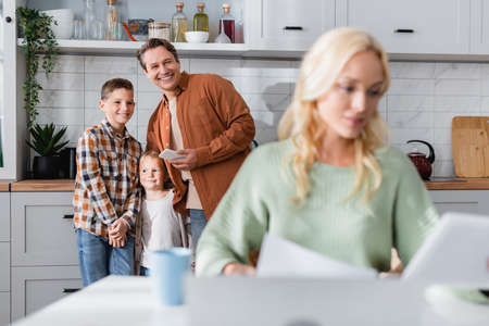 happy man with kids near wife working in kitchen on blurred foregroundの写真素材