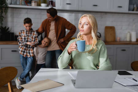 thoughtful woman with cup of tea sitting near laptop, documents, and blurred husband with kids in kitchenの写真素材