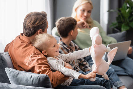 cheerful playing with toy bunny near blurred parents and brother sitting on couchの写真素材