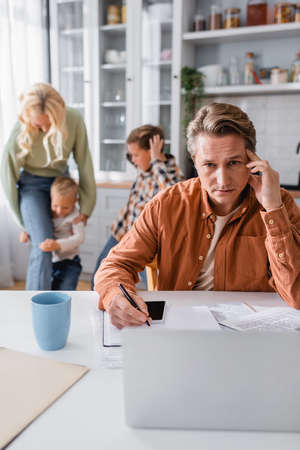 thoughtful man writing in notebook while working in kitchen near mother and sons having fun on blurred backgroundの写真素材