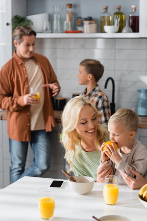 pleased kid eating orange near mom and blurred dad with brother in kitchenの写真素材