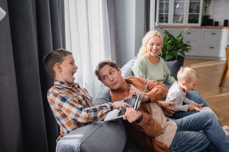 excited boy take away laptop from dad sitting on couch near wife and little kid