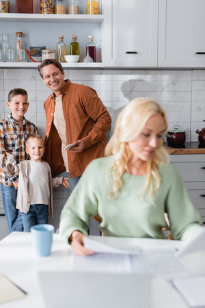 man with son smiling near busy mom working in kitchen near documents and blurred laptopの写真素材
