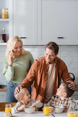 happy dad touching heads of sons having breakfast near mom talking on mobile phone