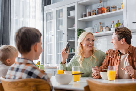 joyful woman with mobile phone looking at husband during breakfast with blurred kidsの写真素材