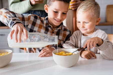 kid pouring milk into bowl with tasty corn flakes while having breakfast with brotherの写真素材