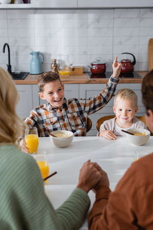 happy boy pointing with finger during breakfast with brother and parents holding hands on blurred foregroundの写真素材