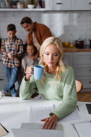 woman with cup of tea working near documents and laptop near blurred family in kitchenの写真素材