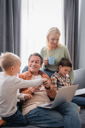 happy woman with cup of tea near husband with laptop having fun with kids on sofa