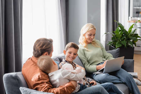 smiling woman with laptop working on sofa near cheerful sons and husbandの写真素材