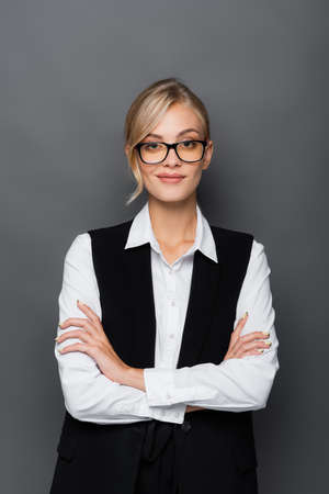 Smiling businesswoman in eyeglasses standing with crossed arms isolated on grey
