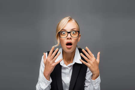 Shocked businesswoman in eyeglasses looking at camera isolated on grey