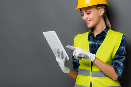 Smiling blonde builder in safety vest and gloves pointing at digital tablet isolated on grey
