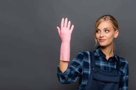 Smiling blonde plumber looking at hand in rubber glove isolated on grey