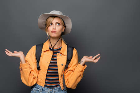 Pensive tourist in hat showing shrug gesture isolated on grey
