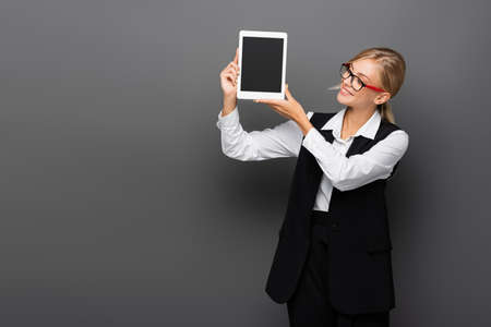 Smiling businesswoman in eyeglasses holding digital tablet with blank screen on grey