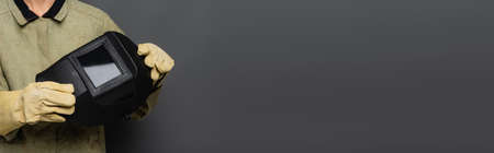 Cropped view of welder in gloves holding helmet with visor on grey background, banner
