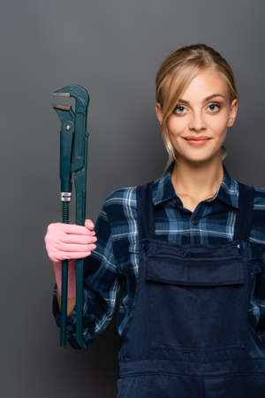 Young blonde plumber in plaid shirt and overalls holding pipe wrench on grey backgroundの写真素材