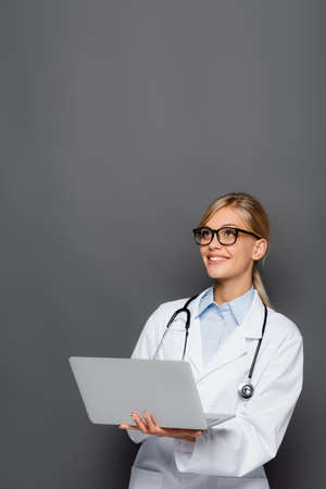 Smiling doctor in eyeglasses holding laptop and looking away isolated on greyの写真素材