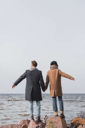 back view of young couple in autumn coats holding hands while standing on sea coastの写真素材