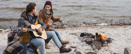 happy woman holding thermo near man playing guitar while sitting on sea coast near bonfire, bannerの写真素材