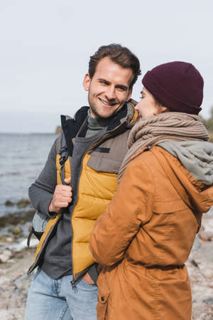 smiling man in autumn clothes looking at girlfriend while walking near river