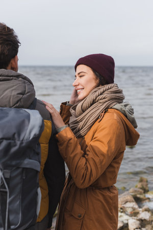 young woman smiling while touching shoulder of man with backpack near riverの写真素材