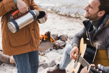 happy man playing guitar near woman pouring drink from thermoの写真素材