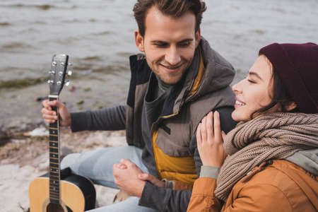 joyful couple in autumn clothes looking at each other while sitting on sea shoreの写真素材