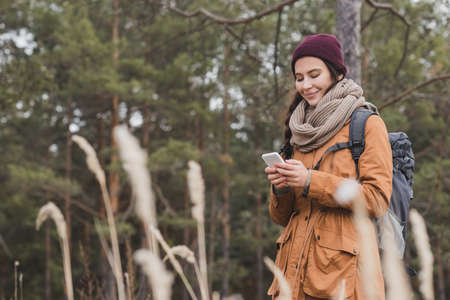 happy woman in autumn outfit using mobile phone during walk in forestの写真素材