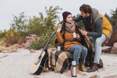 young woman sitting with thermo cup near thermo and guitar while boyfriend covering her with warm blanketの写真素材