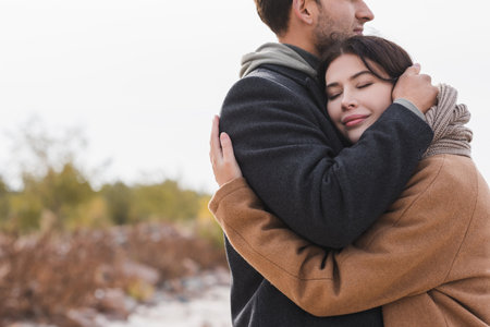 pleased woman with closed eyes hugging man in autumn coat outdoorsの写真素材