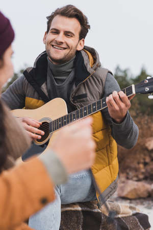 happy young man in autumn outfit playing guitar to blurred woman during autumn walkの写真素材