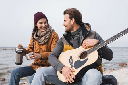 cheerful woman with thermos and man with guitar sitting at riverside during autumn walkの写真素材