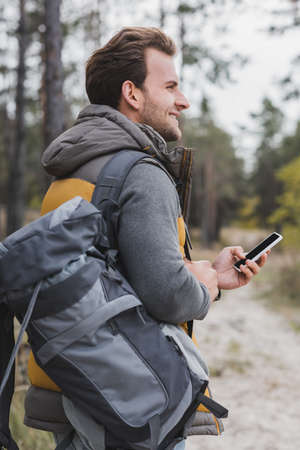 cheerful man with backpack and mobile phone holding cellphone while looking away in forestの写真素材