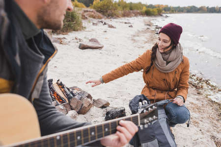 blurred man playing guitar near smiling woman warming near bonfireの写真素材