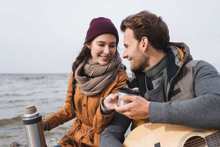 smiling man taking warm drink from woman with thermo while sitting near seaの写真素材