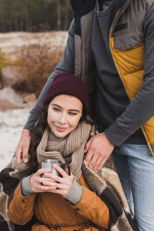 smiling woman holding thermo cup near man covering her with plaid blanket outdoorsの写真素材
