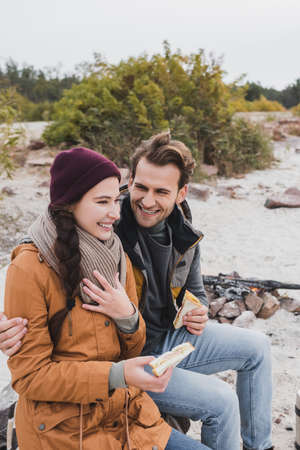 joyful couple holding sandwiches while sitting near bonfire during haltの写真素材