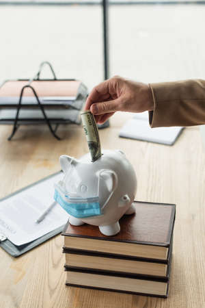 partial view of lawyer putting dollar banknote into piggy bank in medical mask, anti-corruption...