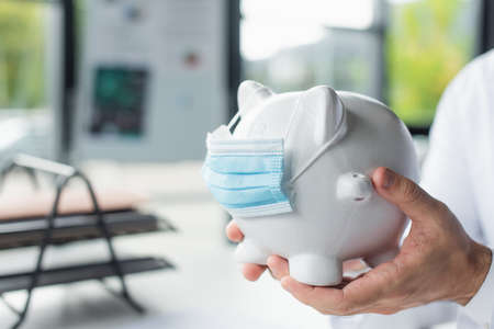 cropped view of doctor holding piggy bank in medical mask in hospital, anti-corruption concept