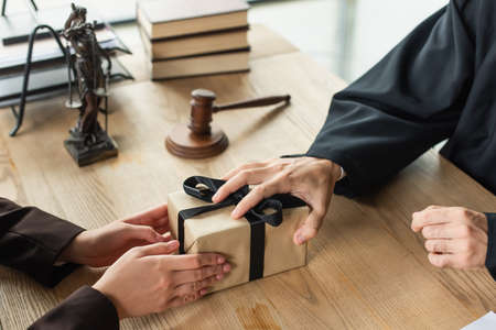 cropped view of judge taking gift from client near blurred gavel on desk, anti-corruption concept