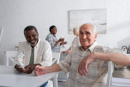 Elderly man looking at camera near smiling african american friend with smartphone in nursing home