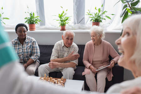 Senior interracial people with digital tablet talking near chess and blurred nurse in nursing home